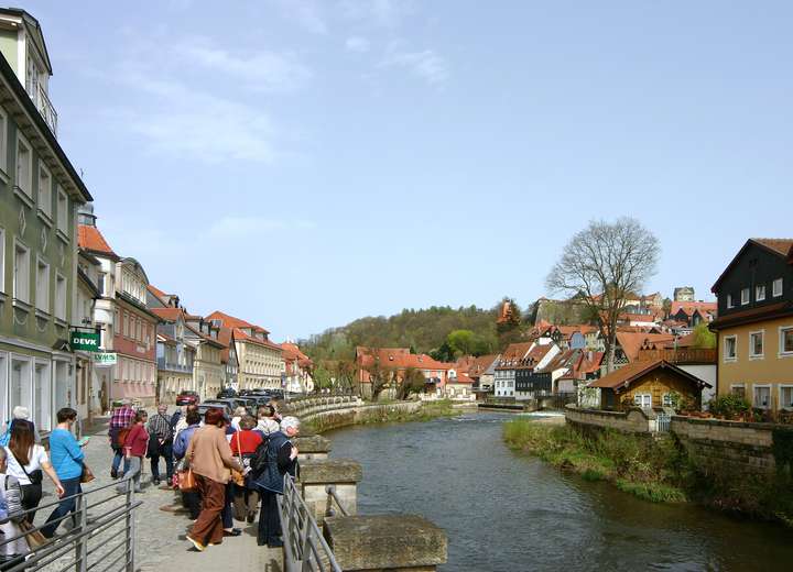 Menschengruppe schlendert entlang des Flüsses Haßlach, Häuser reihen sich links und rechts vom Ufer an