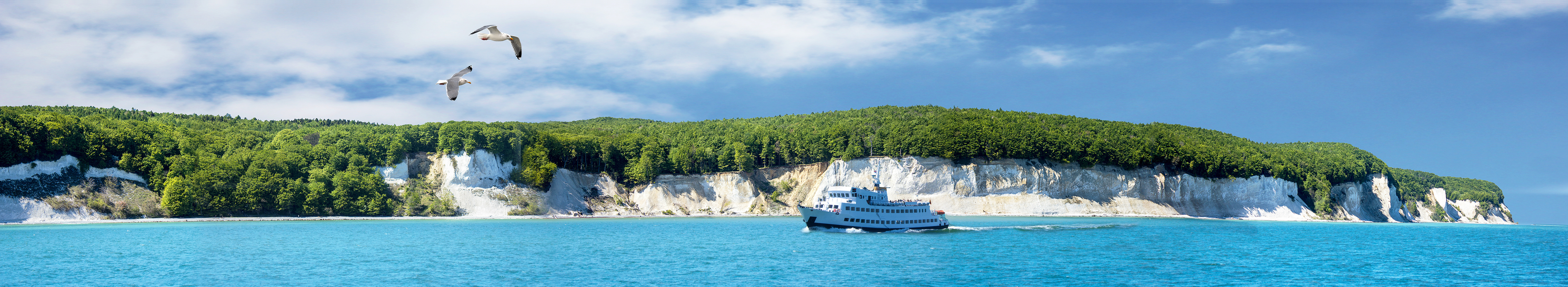 Weißer Kreidefelsen an der Küste von Rügen im Jasmund Nationalpark mit einem Boot auf dem blauen Meer und Möwen am Himmel.